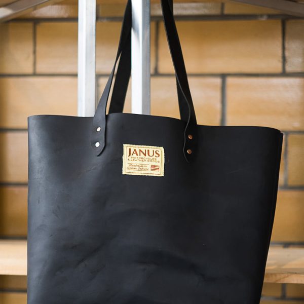 Black leather tote bag with JANUS logo hanging on a bench against brick wall background.