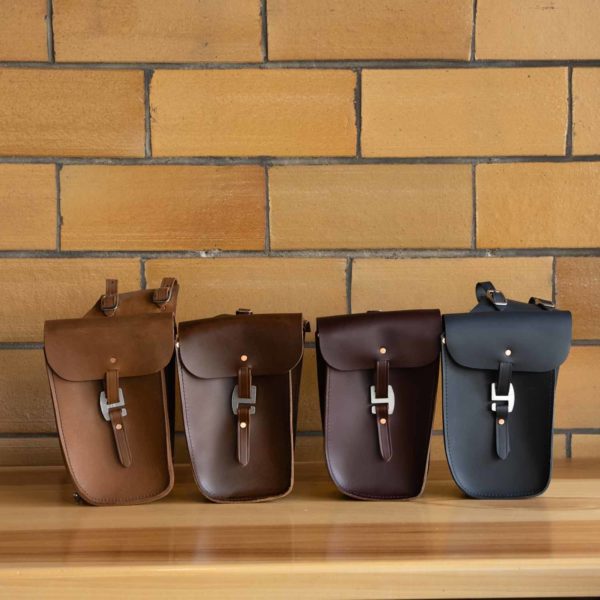 Four leather saddlebags in varying shades on a wooden shelf against a brick wall.