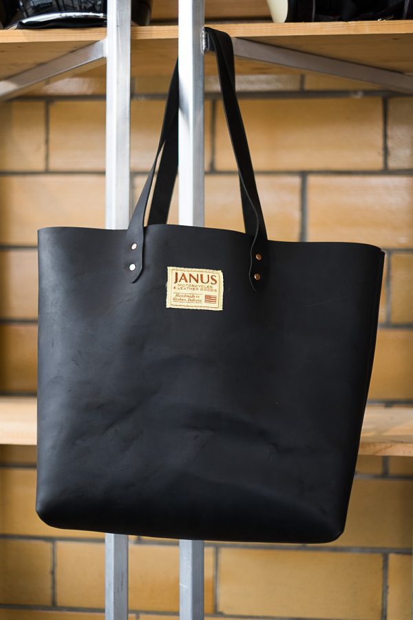 Black leather tote bag with JANUS logo hanging on metal rack.