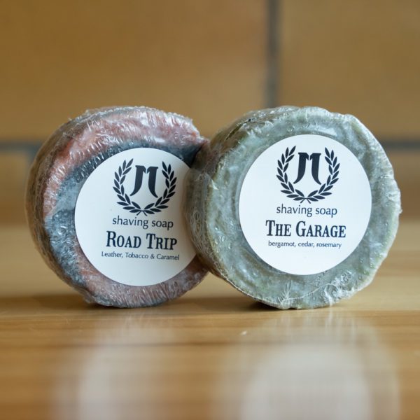 Two artisan shaving soaps labeled Road Trip and The Garage on a wooden surface.