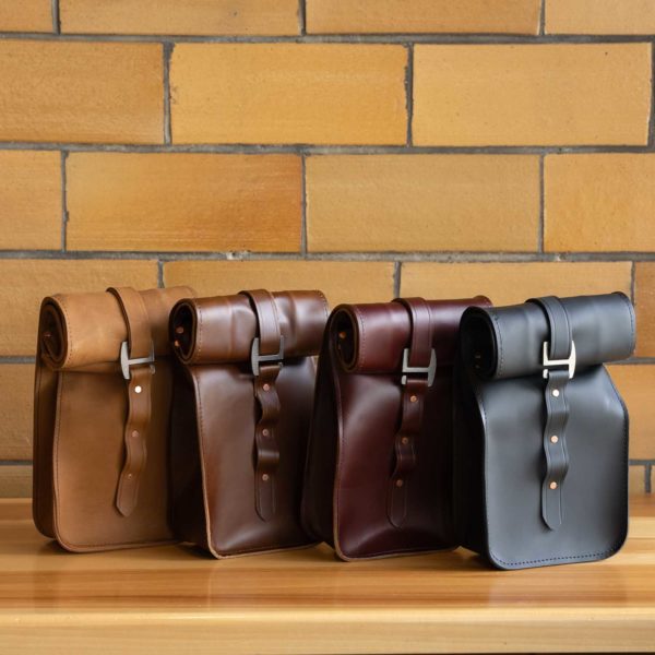 Three leather messenger bags in tan, brown, and black on a wooden shelf against a brick wall.