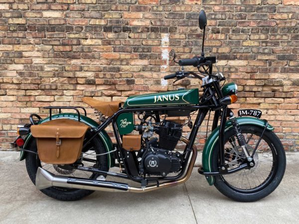 Green Janus motorcycle with leather saddlebags parked against brick wall.