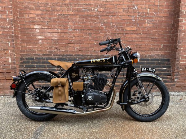 Black Janus motorcycle with leather saddlebags parked against a brick wall.