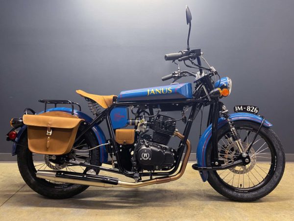 Blue Janus motorcycle with leather saddlebags parked against a gray wall.