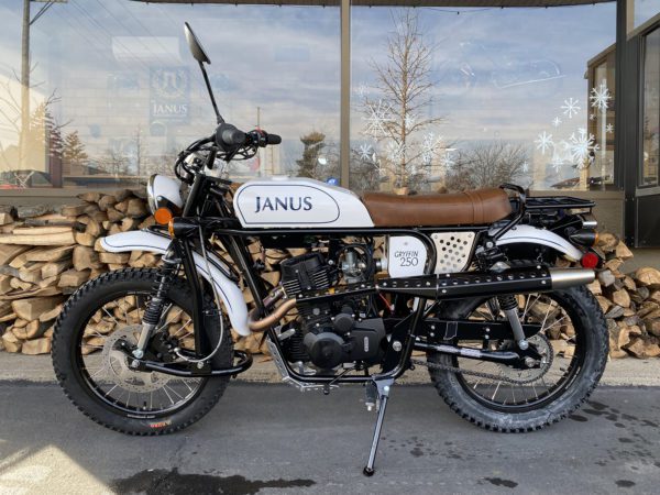 White and black Janus Gryffin 250 motorcycle parked in front of a woodpile and glass storefront.