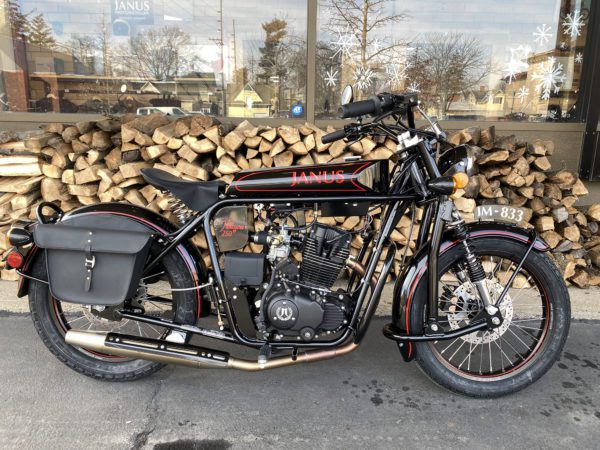 Black retro Janus motorcycle parked in front of a woodpile.