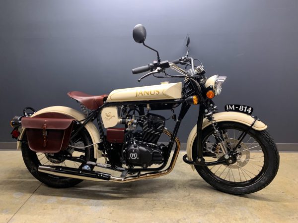Classic cream-colored Janus motorcycle with dark leather saddlebags on concrete floor.