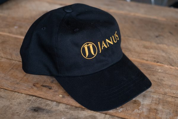 Black baseball cap with gold 'JANUS' embroidery on wooden surface.