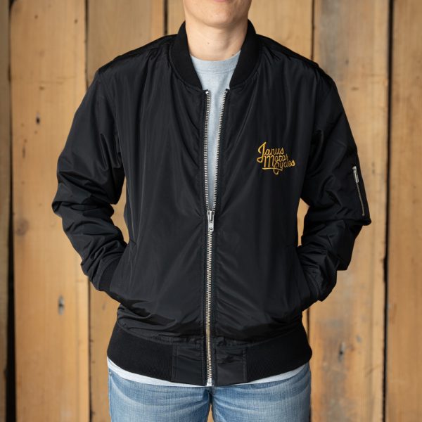 Person wearing a black bomber jacket with logo, standing against a wooden background.