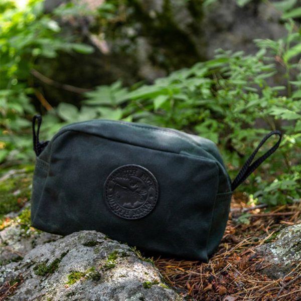 Green toiletry bag with logo on forest floor among mossy rocks.